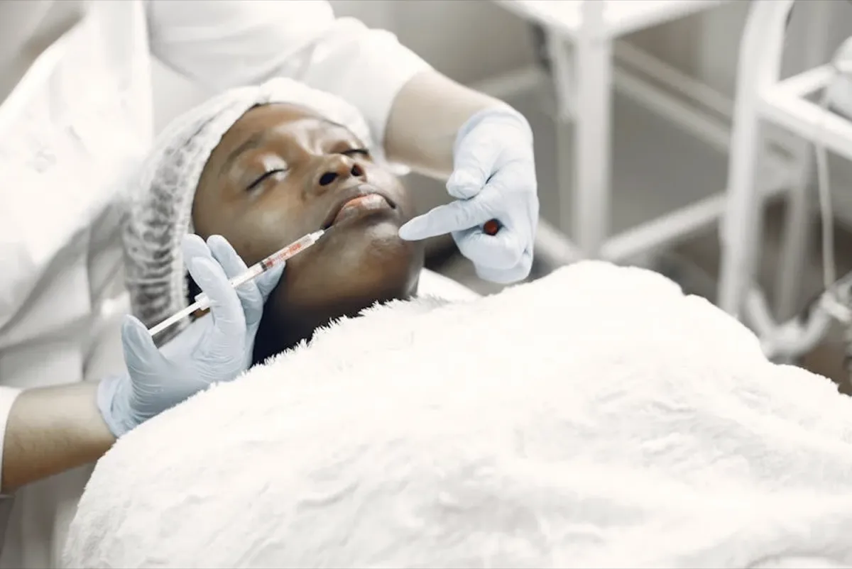 A gloved clinician injecting filler into a reclining patient’s lower lip.