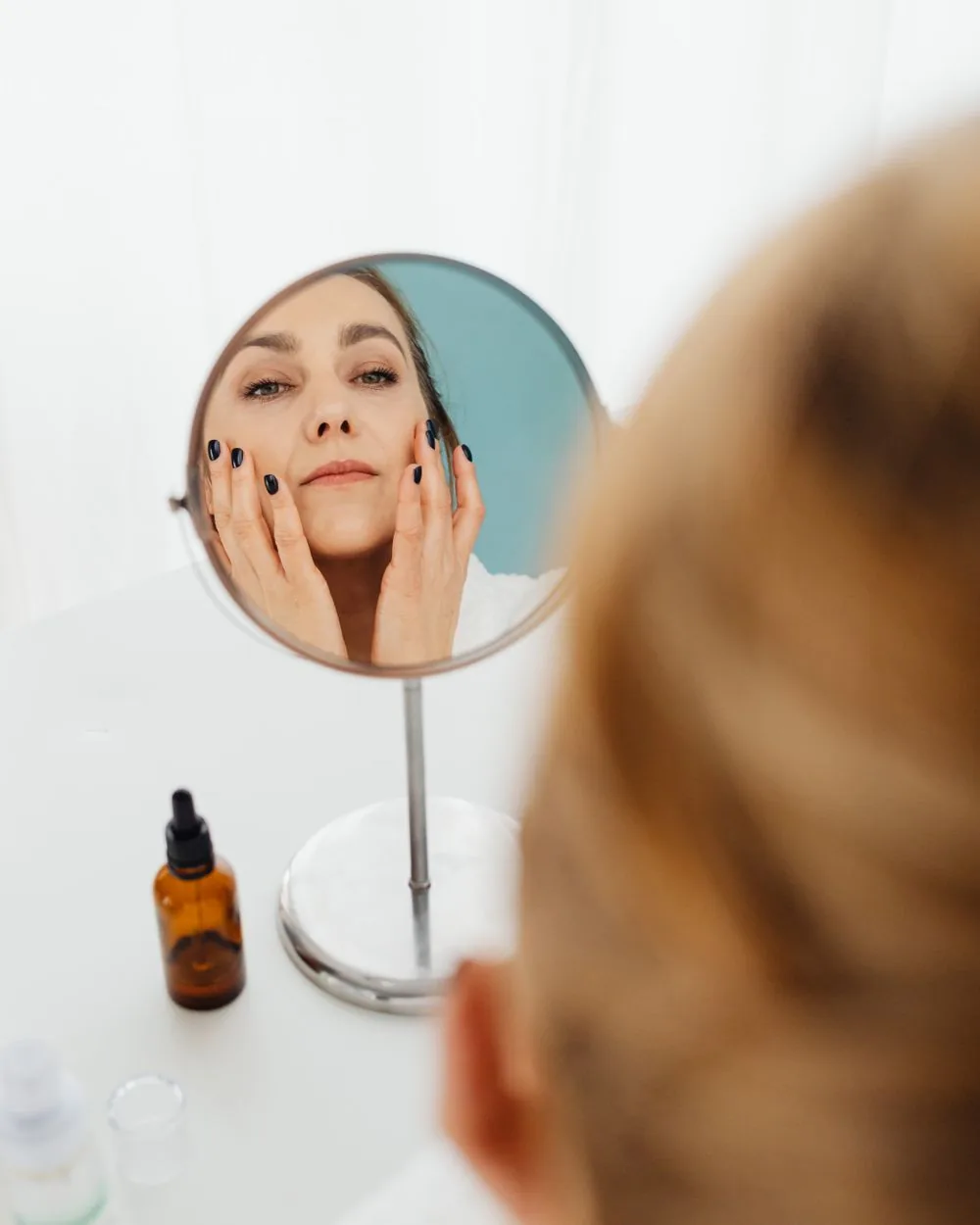 A woman examining her face in a mirror, hands resting on her cheeks.