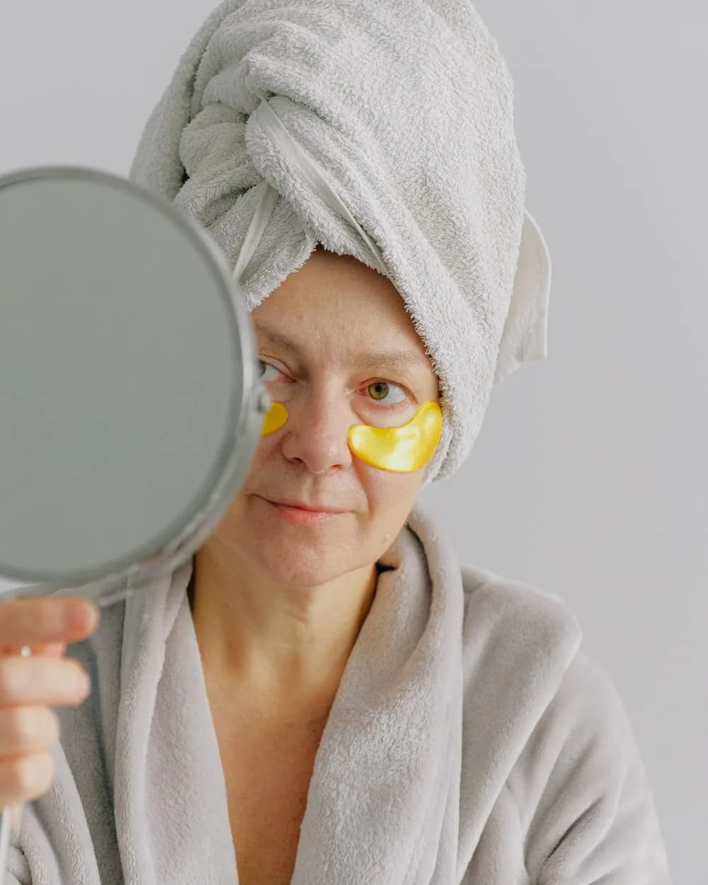 A woman holding a mirror, wearing under-eye masks.